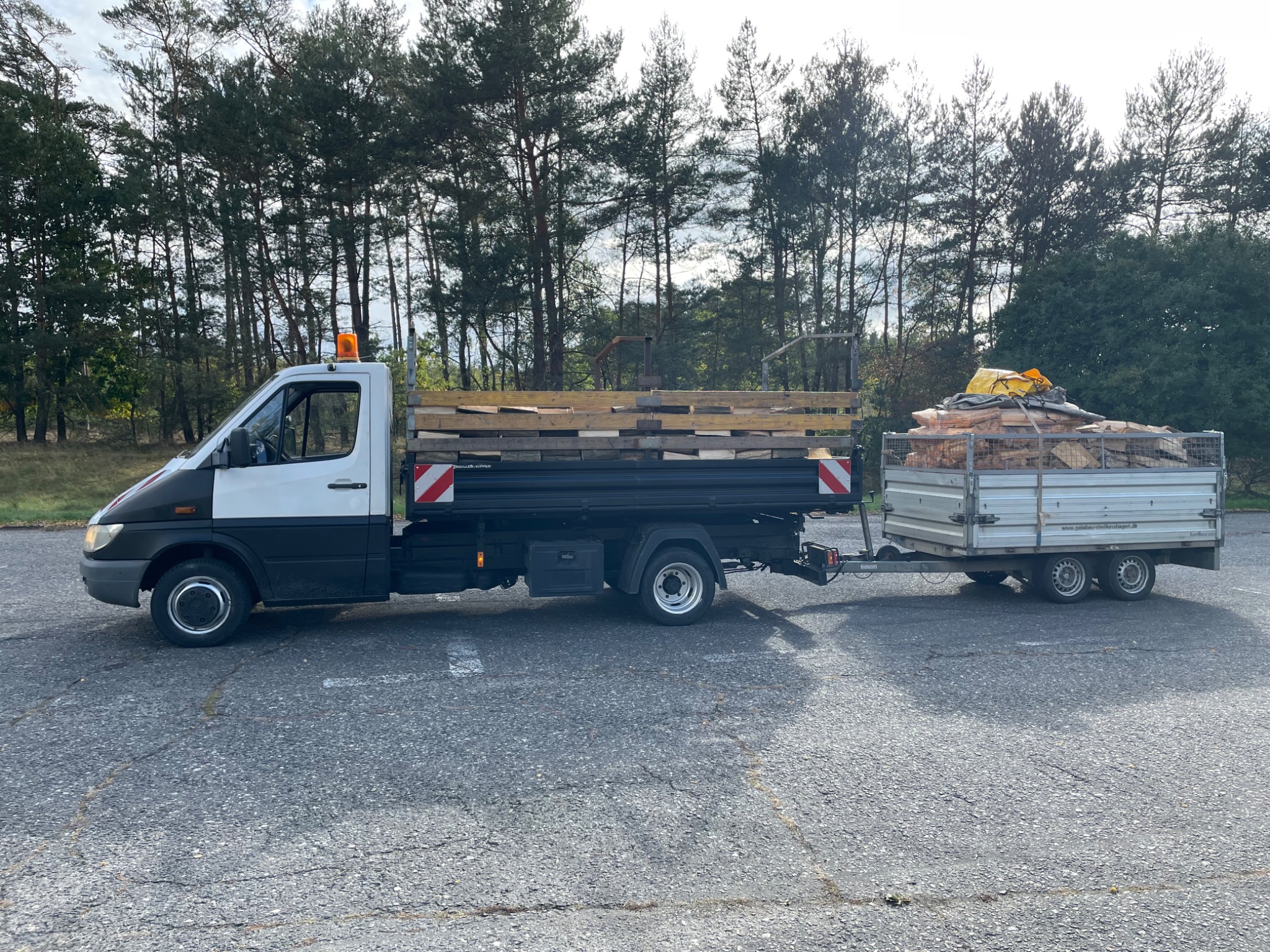 Ciężarówka z przyczepą wypełniona drewnem na tle drzew, widok z boku na szarym asfalcie.