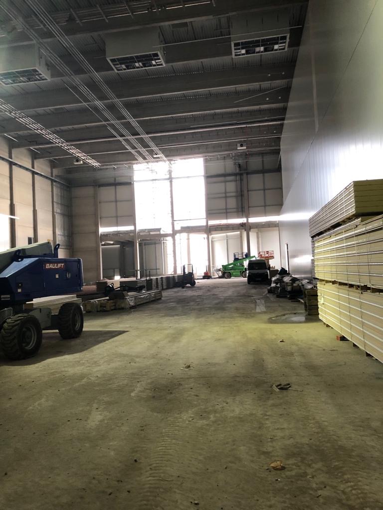 Wnętrze hali w trakcie budowy z widocznymi płytami warstwowymi ułożonymi przy ścianie, podnośnikami koszowymi i przygotowanymi elementami konstrukcyjnymi.