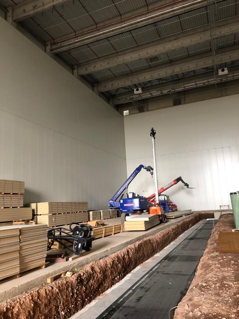 Wnętrze hali w trakcie budowy z widocznymi płytami warstwowymi, podnośnikami teleskopowymi i wykopem wzdłuż ściany.