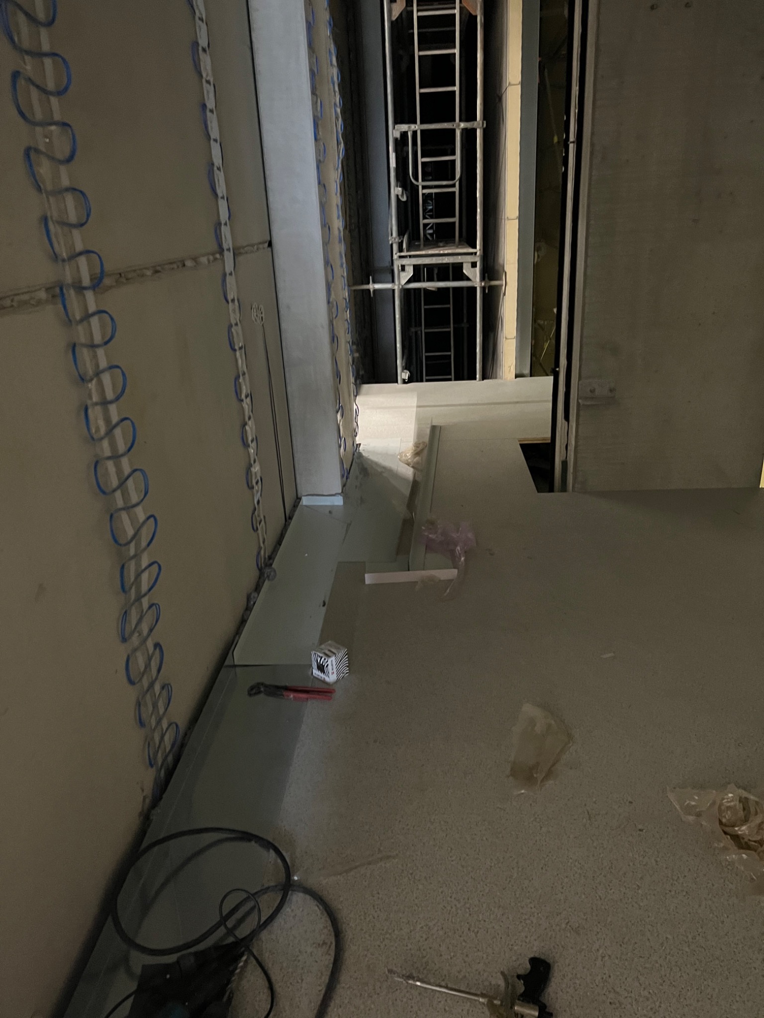 Wnętrze hali w trakcie budowy, widoczne płyty warstwowe z niebieskimi elementami mocującymi, rusztowanie w głębi i narzędzia na podłodze.