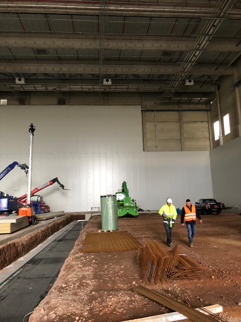 Wnętrze hali w trakcie budowy, widoczne płyty warstwowe na ścianach, pracownicy w odblaskowych kamizelkach, maszyny budowlane i przygotowane zbrojenia.