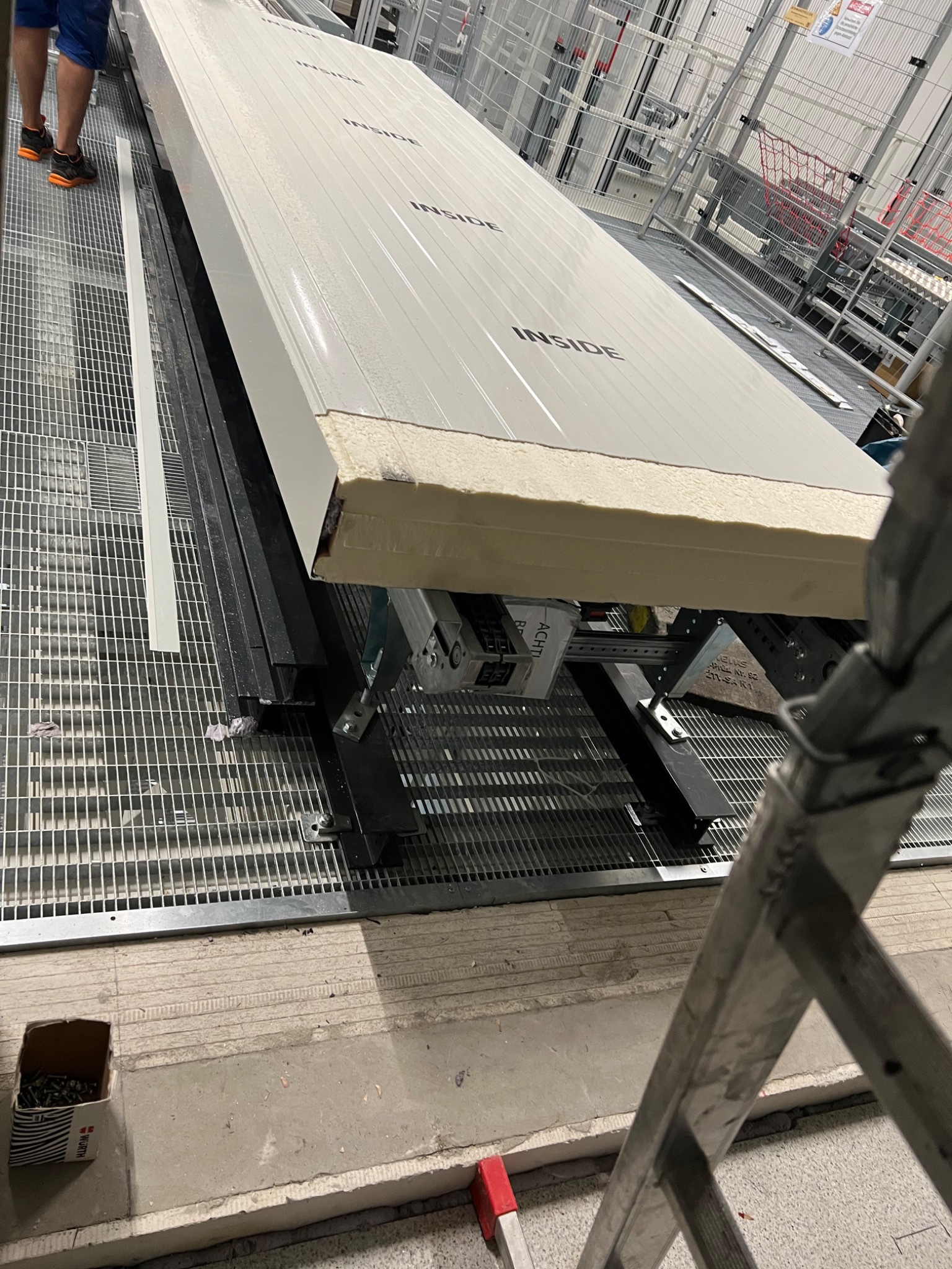 Widok z dołu na przygotowaną płytę warstwową z napisem 'INSIDE' leżącą na metalowej konstrukcji, widoczna warstwa izolacyjna, w tle rusztowania i elementy konstrukcyjne hali, na pierwszym planie...