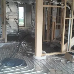 Instalacja ogrzewania podłogowego w trakcie montażu, rury ułożone na folii, widoczne elementy konstrukcji drewnianej i izolacji ścian. W tle okno z widokiem na zewnątrz.