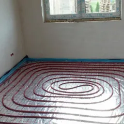 Instalacja ogrzewania podłogowego z czerwonymi rurami na folii refleksyjnej w surowym pomieszczeniu z oknami.