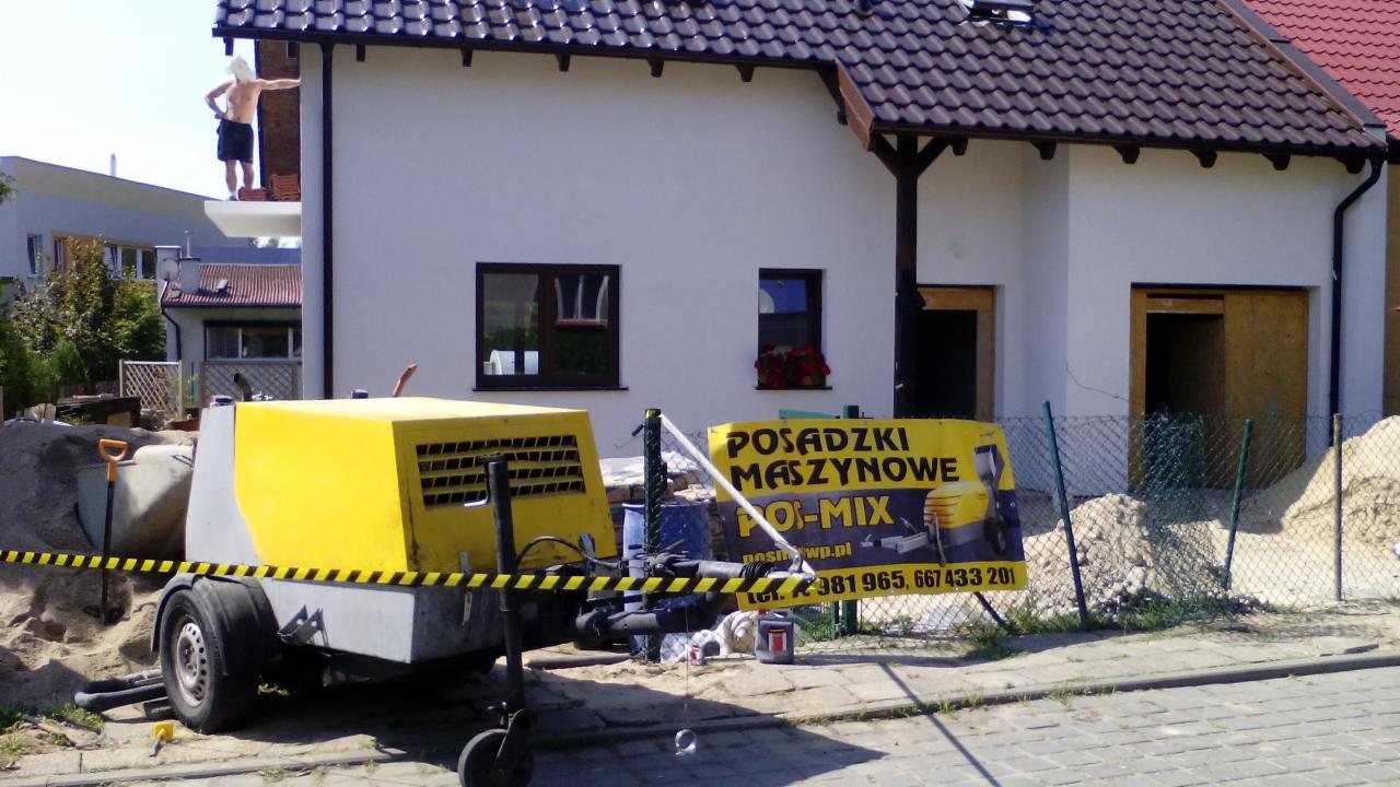 Żółto-szara maszyna do posadzek maszynowych PO-MIX przed budynkiem z oknami i robotnikiem na dachu. Żółto-czarna taśma ostrzegawcza. Reklama firmy w tle.