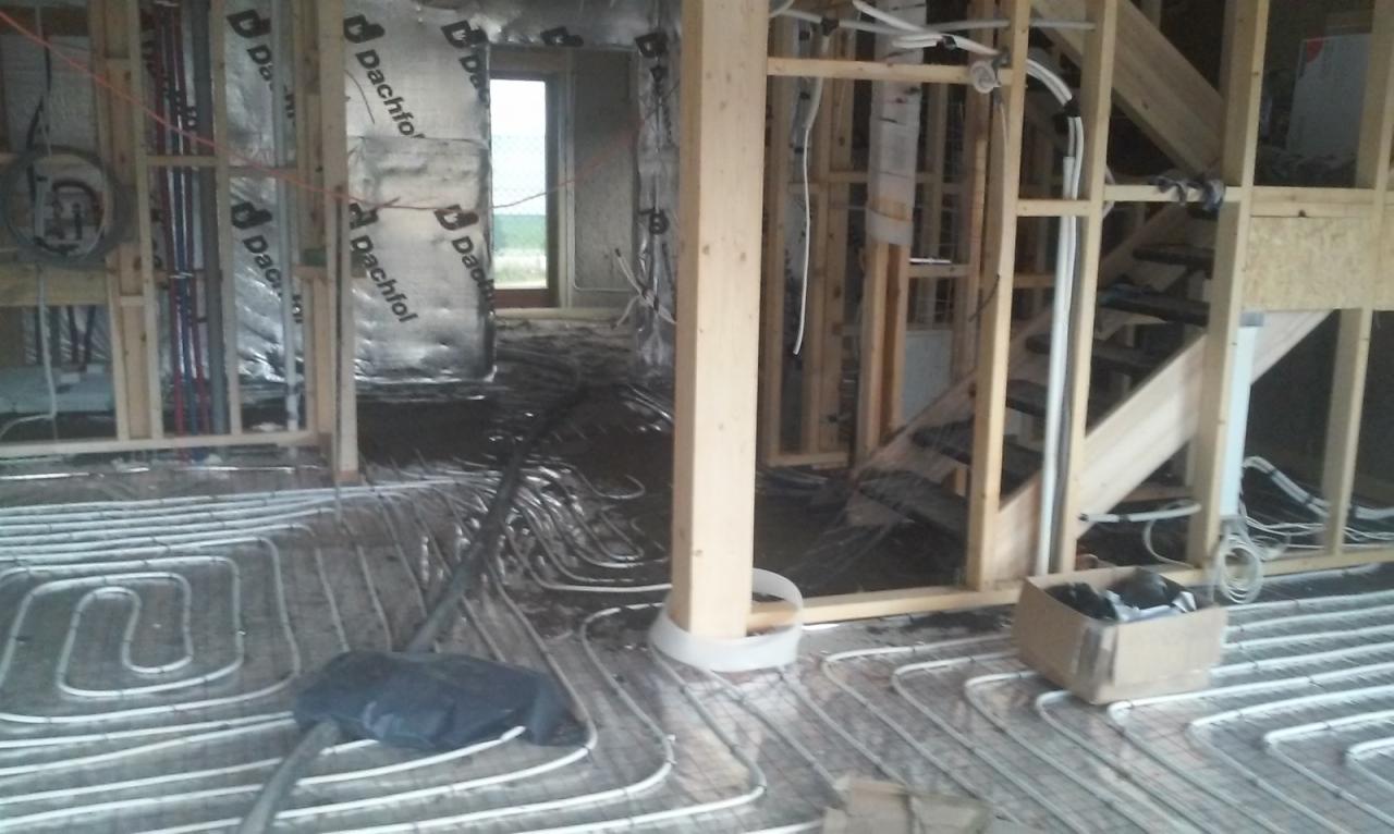 Instalacja ogrzewania podłogowego w trakcie montażu, rury ułożone na folii, widoczne elementy konstrukcji drewnianej i izolacji ścian. W tle okno z widokiem na zewnątrz.