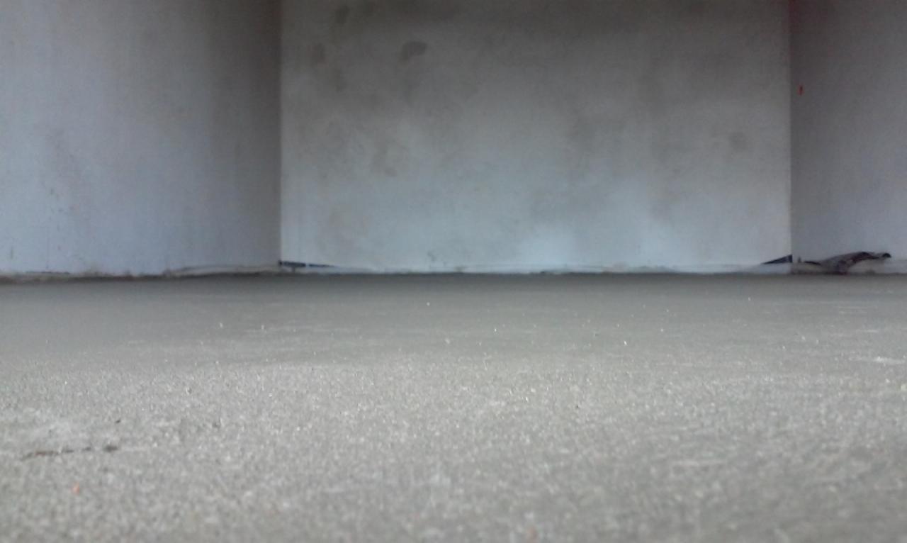 Świeżo wylana, gładka posadzka w szarym odcieniu, widok z niskiej perspektywy, surowe ściany w tle. Widoczne czarne elementy uszczelniające przy ścianach.