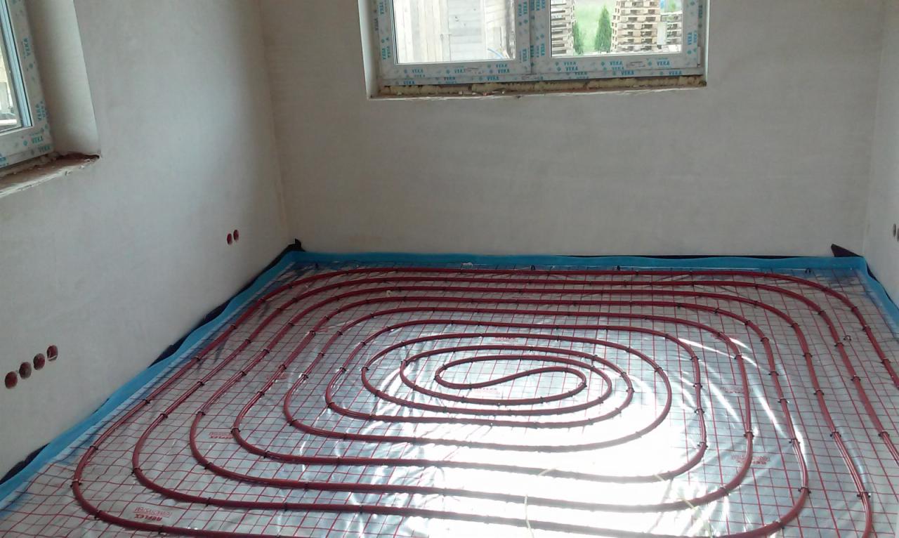 Instalacja ogrzewania podłogowego z czerwonymi rurami na folii refleksyjnej w surowym pomieszczeniu z oknami.