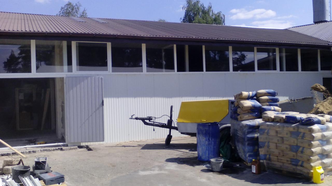 Budynek przemysłowy z brązowym dachem, białą elewacją i rzędem okien. Przed budynkiem worki z zaprawą, przyczepka i narzędzia budowlane.