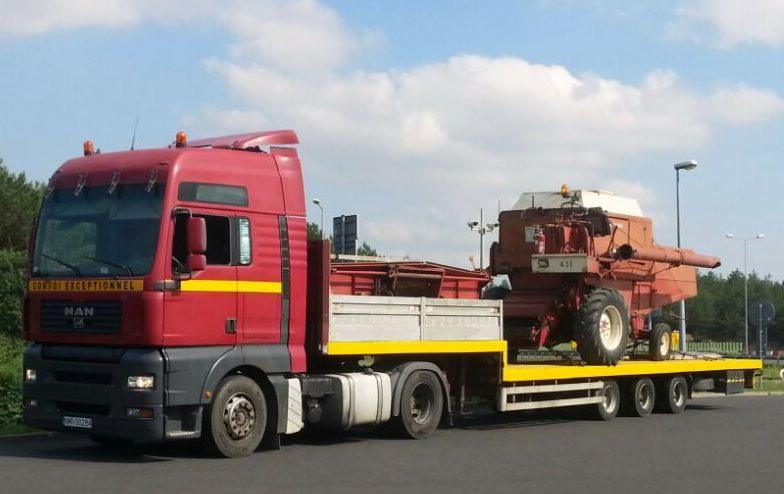 Czerwony ciągnik siodłowy MAN przewozi na żółtej platformie kombajn rolniczy w słoneczny dzień. Na kabinie widoczny napis 'Convoi Exceptionnel'.