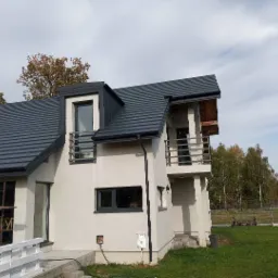 Nowoczesny dom z ciemnoszarą dachówką ceramiczną, widoczna obróbka blacharska, balkon z metalową balustradą, częściowo widoczne rusztowanie przy elewacji.