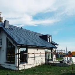 Nowoczesny dom jednorodzinny z grafitowym dachem z dachówki ceramicznej, widoczne rusztowania wokół elewacji, błękitne niebo w tle.