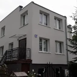 Dwupiętrowy dom murowany z szarą elewacją, czarną balustradą schodów wejściowych i numerem '2' na ścianie.