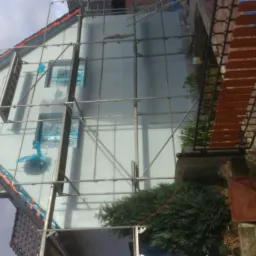 Dom jednorodzinny w trakcie prac elewacyjnych, zabezpieczone okna folią, rusztowanie metalowe przy ścianie, widoczny fragment dachu z dachówką.