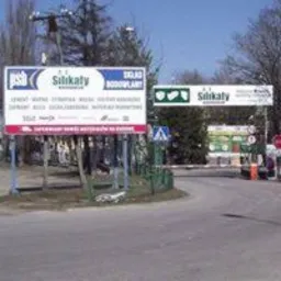 Tablica reklamowa z napisem 'Skład Budowlany' i logo 'Silikaty' przy wjeździe na teren firmy, widoczne materiały budowlane w tle.