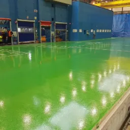 Świeżo wylana, jasnozielona posadzka w hali przemysłowej z białymi liniami wyznaczającymi pola, widoczne otwarte bramy i pracownicy w oddali oraz niebieska ściana w tle.