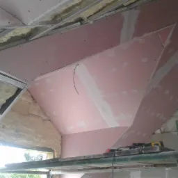 Montaż różowych płyt kartonowo-gipsowych na poddaszu, widoczne metalowe profile konstrukcyjne, izolacja z wełny mineralnej oraz narzędzia na rusztowaniu.