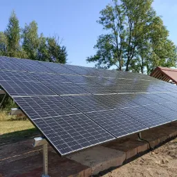 Instalacja fotowoltaiczna w Mniszkowie, widok z dołu na panele słoneczne zamontowane na metalowych wspornikach, z ceglaną podstawą i zielenią w tle, pod błękitnym niebem.