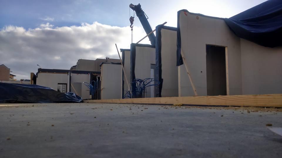 Wznoszenie domu modułowego: fragment konstrukcji z widocznym dźwigiem na tle pochmurnego nieba. Elementy domu w trakcie montażu, zabezpieczone folią.