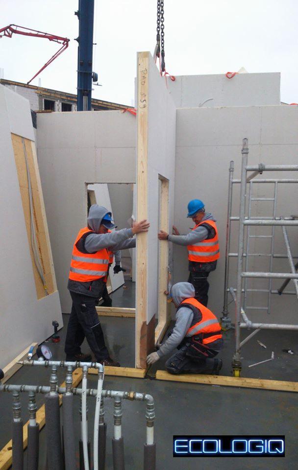 Montaż ściany modułowej z oknem przez trzech pracowników w kamizelkach, widoczny dźwig, rury instalacji i rusztowanie. Budowa w toku, surowy stan.