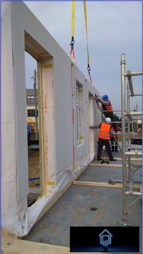 Montaż ściany modułowej domu z oknami przez dwóch pracowników w kaskach i kamizelkach. Ściana podwieszona na pasach, widoczne rusztowania i elementy konstrukcyjne.