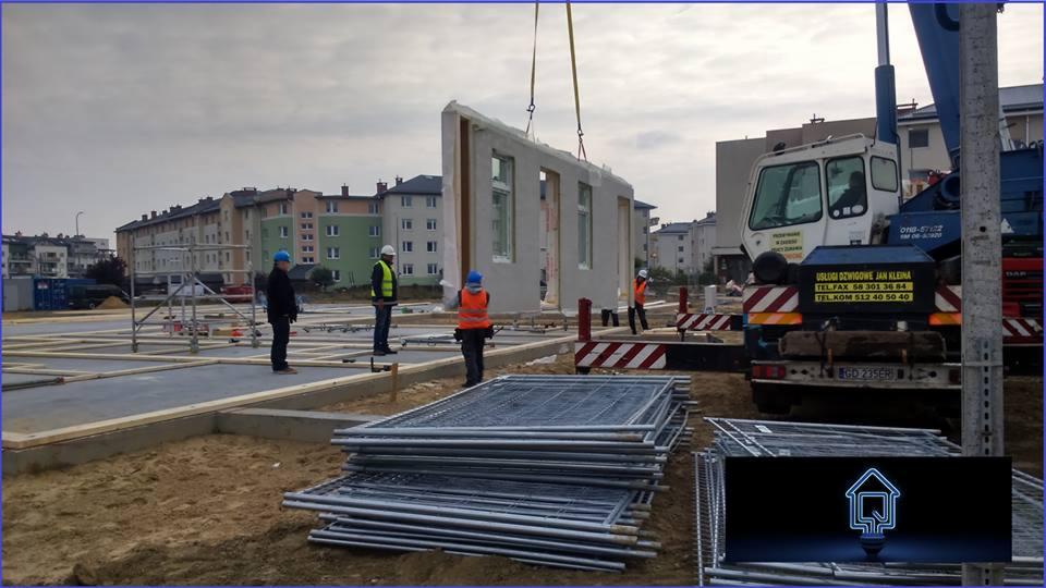 Montaż ściany domu modułowego z oknami za pomocą dźwigu. Widoczni pracownicy w kaskach i kamizelkach odblaskowych. W tle zabudowa miejska i elementy szalunku.