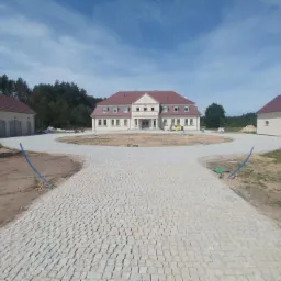 Szeroki kadr na podjazd do dużego domu z dwoma budynkami gospodarczymi, wyłożony jasną kostką, z widocznymi niebieskimi rurami na obrzeżach i śladami prac ziemnych.