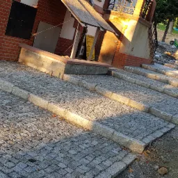 Kamienne stopnie schodów zewnętrznych w trakcie budowy, widoczne nierówności terenu i materiały budowlane, ciepłe światło słoneczne podkreśla fakturę kamienia.