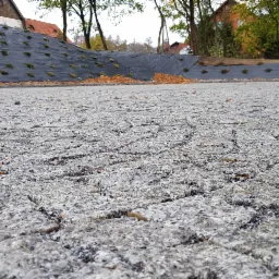 Szara nawierzchnia z kostki, z widocznym w tle zboczem zabezpieczonym czarną agrowłókniną z nasadzeniami.