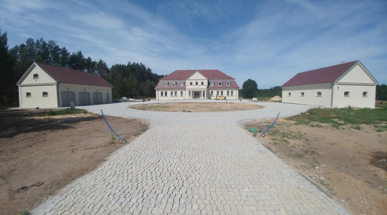 Szeroki kadr na podjazd do dużego domu z dwoma budynkami gospodarczymi, wyłożony jasną kostką, z widocznymi niebieskimi rurami na obrzeżach i śladami prac ziemnych.