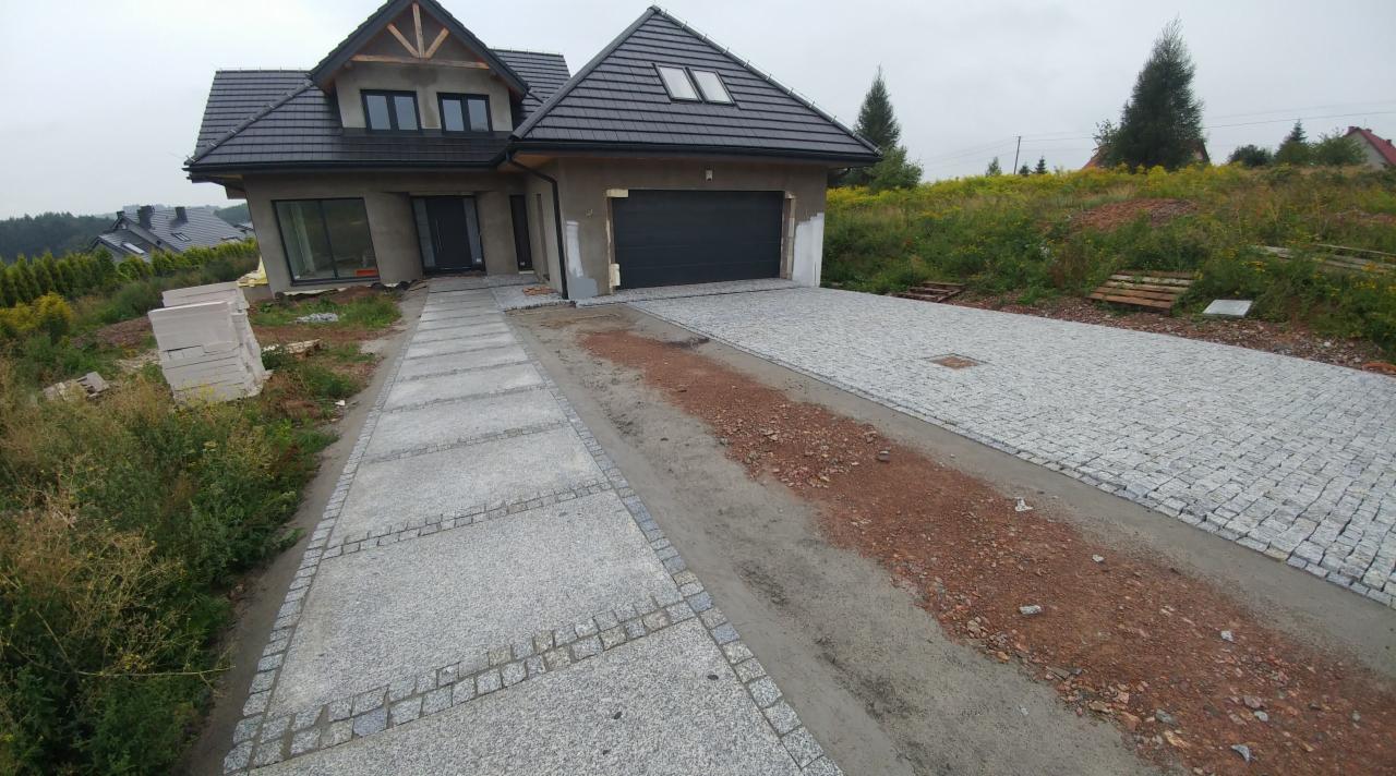 Ułożona nawierzchnia z kostki granitowej prowadząca do garażu nowoczesnego domu jednorodzinnego na wzgórzu, widoczny fragment podjazdu z podsypką i paleta kostki obok domu.