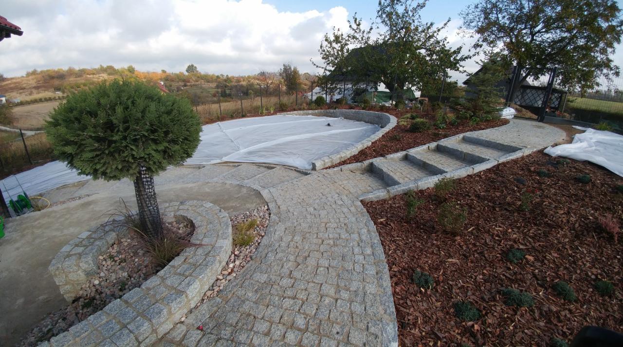 Ogród z ścieżką z kostki, schodami i okrągłym rabatem z drzewem. W tle zadaszony basen i dom.