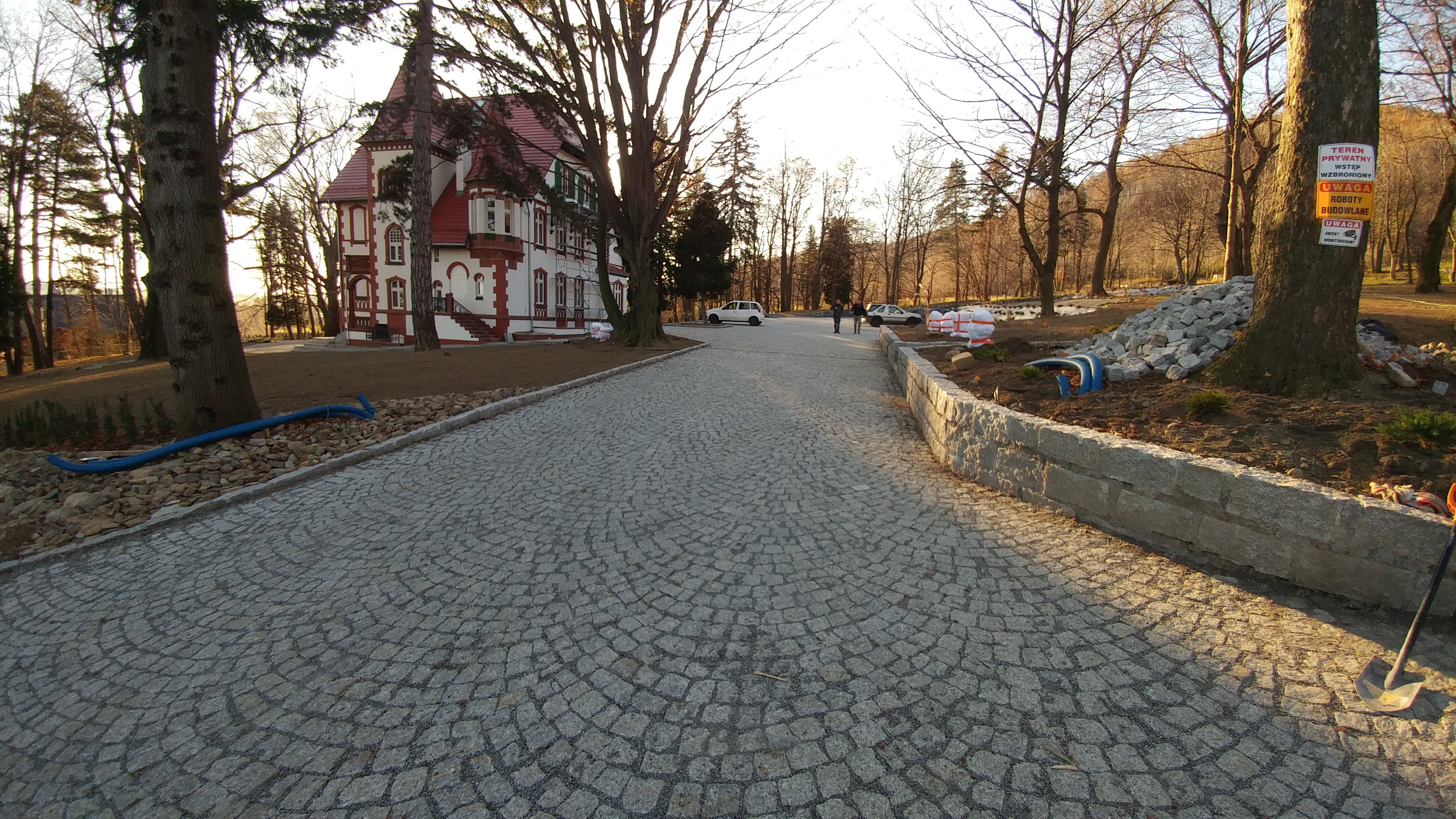 Szeroki kadr na podjazd z szarej kostki ułożonej w wachlarz, z widocznym budynkiem w stylu dworku i przygotowanymi rabatami po bokach, w tle drzewa i tabliczka informująca o robotach budowlanych.