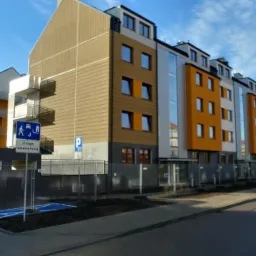 Nowoczesny budynek wielorodzinny z balkonami, wykończony w kolorach białym, żółtym i brązowym, widoczny znak drogowy 'Droga wewnętrzna' i miejsce parkingowe dla niepełnosprawnych, teren ogrodzony...