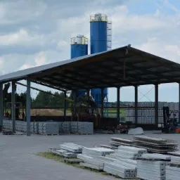 Otwarta hala magazynowa z prefabrykatami betonowymi i silosami w tle. Wózek widłowy transportuje materiały. Widok na plac składowy z elementami konstrukcyjnymi.