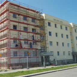 Elewacja budynku w trakcie prac. Na rusztowaniach ekipa malarzy w kombinezonach. Część budynku w kolorze kremowym, druga w trakcie wykonywania.