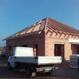 Budowa parterowego budynku z czerwonej cegły z drewnianą konstrukcją dachu, na pierwszym planie biały samochód dostawczy z widoczną tablicą rejestracyjną, w tle inne budynki.