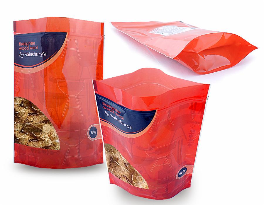 Trzy foliowe torebki stojące z zamknięciem strunowym, z okienkiem pokazującym zawartość - wełnę drzewną rozpałkową, z nadrukiem w kolorze czerwonym i granatowym, na białym tle.