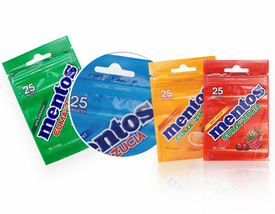 Cztery kolorowe torebki z gumami do żucia Mentos w opakowaniach z zamknięciem strunowym: zielona (spearmint), niebieska, pomarańczowa (cytrusowa) i czerwona (owoce leśne), ustawione pod kątem...