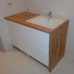 Łazienkowa szafka z białymi frontami i blatem z jasnego drewna, z umywalką wpuszczaną w blat i chromowaną baterią. Widoczne białe płytki na ścianie i podłodze.