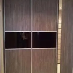 Szafa przesuwna z czterema drzwiami, z których środkowe dwa mają czarne, błyszczące panele, a pozostałe drewnopodobne wykończenie w pionowe prążki.