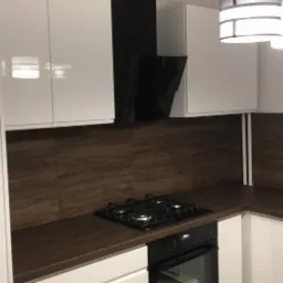 Nowoczesna kuchnia z białymi, lakierowanymi frontami szafek, brązowym blatem i okładziną ścienną imitującą drewno; czarny okap nad płytą gazową i designerska lampa wisząca.