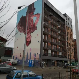 Nowoczesny budynek mieszkalny z ceglaną elewacją i balkonami, na ścianie bocznej mural przedstawiający akordeonistę na tle panoramy miasta.