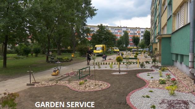 Aranżacja ogrodu przy bloku: rabaty z obrzeżami, kamień ozdobny, świeżo posadzone rośliny. W tle zaparkowane żółte busy. Prace w toku.