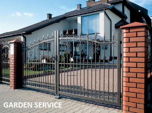 Kuta, metalowa brama wjazdowa z dekoracyjnymi elementami, osadzona na ceglanych słupkach. W tle dom z dużym oknem i zielenią. Całość w słoneczny dzień.