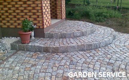 Kamienne schody wejściowe do domu, wyłożone kostką brukową w odcieniach szarości i beżu. Na schodach stoi doniczka z kwiatami, w tle ceglana elewacja i zieleń.