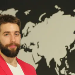 Portret mężczyzny z brodą w czerwonej marynarce na tle mapy świata na ścianie.