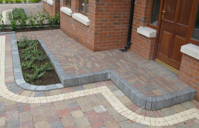 Kaskadowe ułożenie kostki brukowej w odcieniach czerwieni i beżu, obrzeża z szarej kostki, rabata z zielenią przy wejściu do domu z cegły.