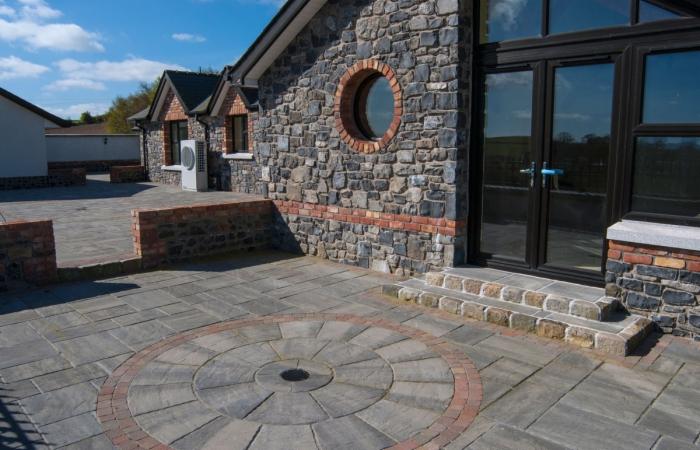 Wykończony taras z kostki brukowej w szarych odcieniach, z okrągłym wzorem i ceglaną obwódką. Kamienna elewacja budynku z okrągłym oknem i czarnymi drzwiami.