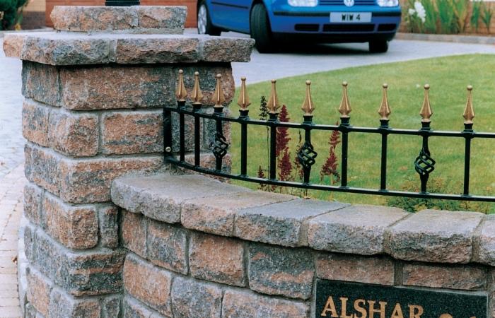 Kamienna ścianka oporowa z czarnym, dekoracyjnym metalowym ogrodzeniem z pozłacanymi elementami, na tle trawnika i częściowo widocznego niebieskiego samochodu.